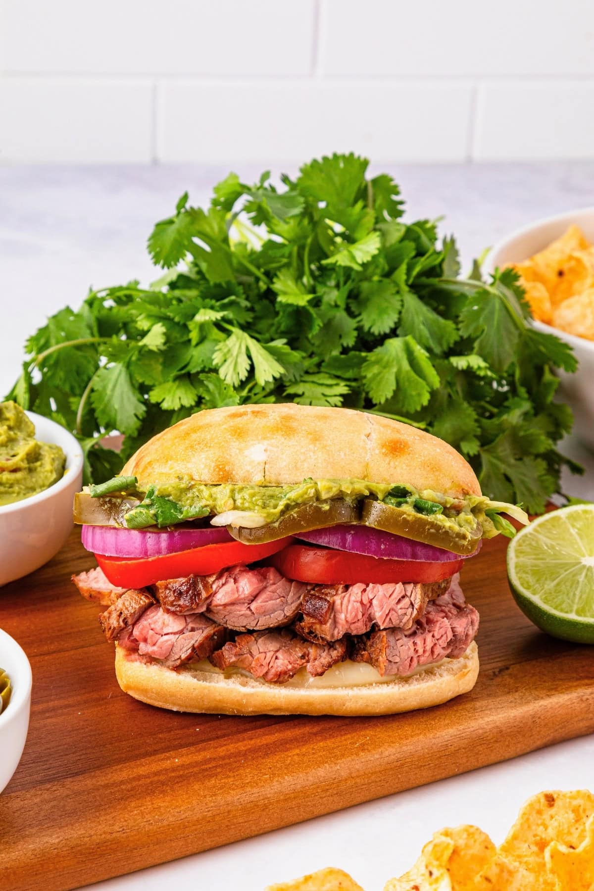 A steak sandwich with tomato, red onion, guacamole, and cilantro on a board.