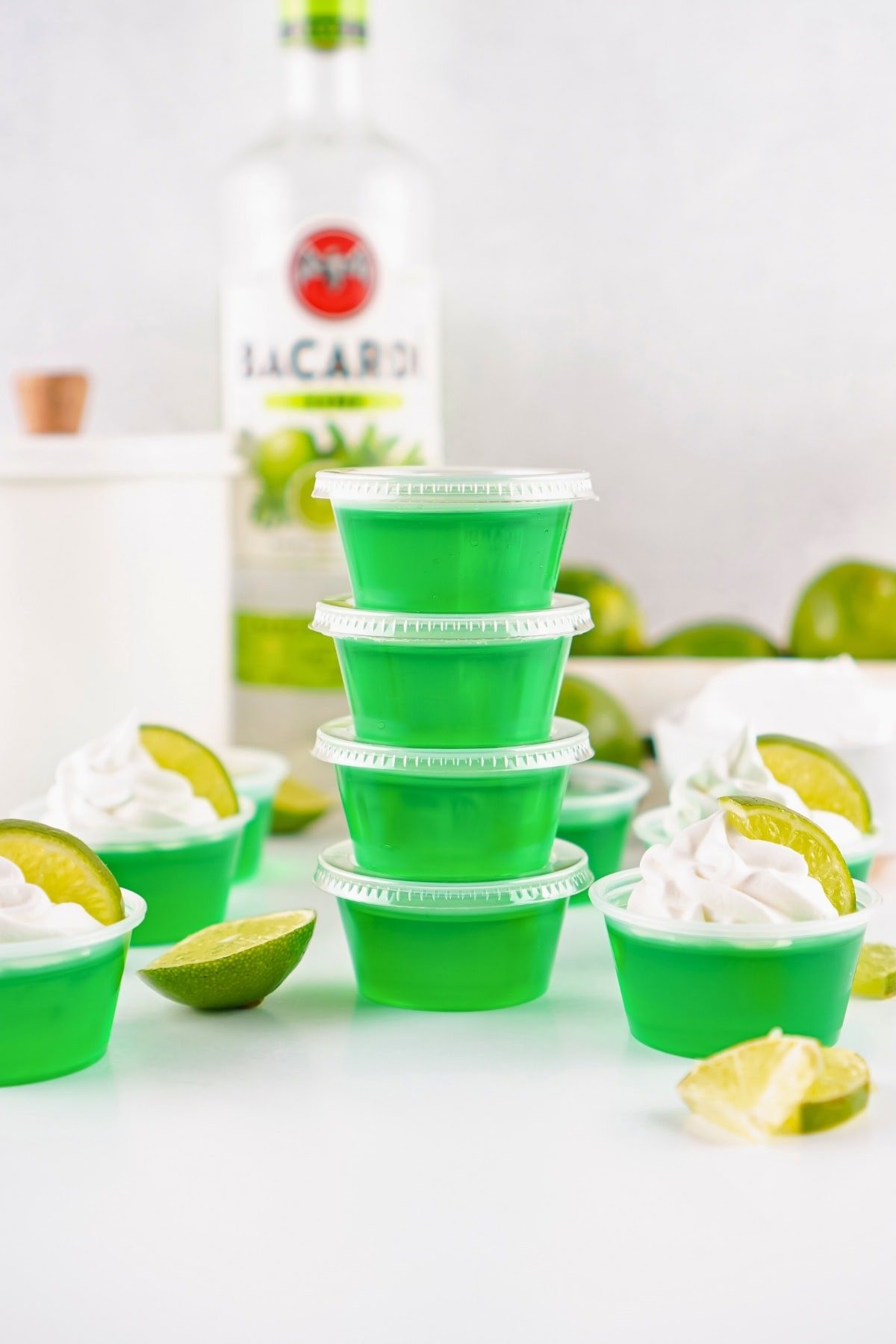 Four stacked green jello shots with limes and whipped cream, Bacardi bottle behind.