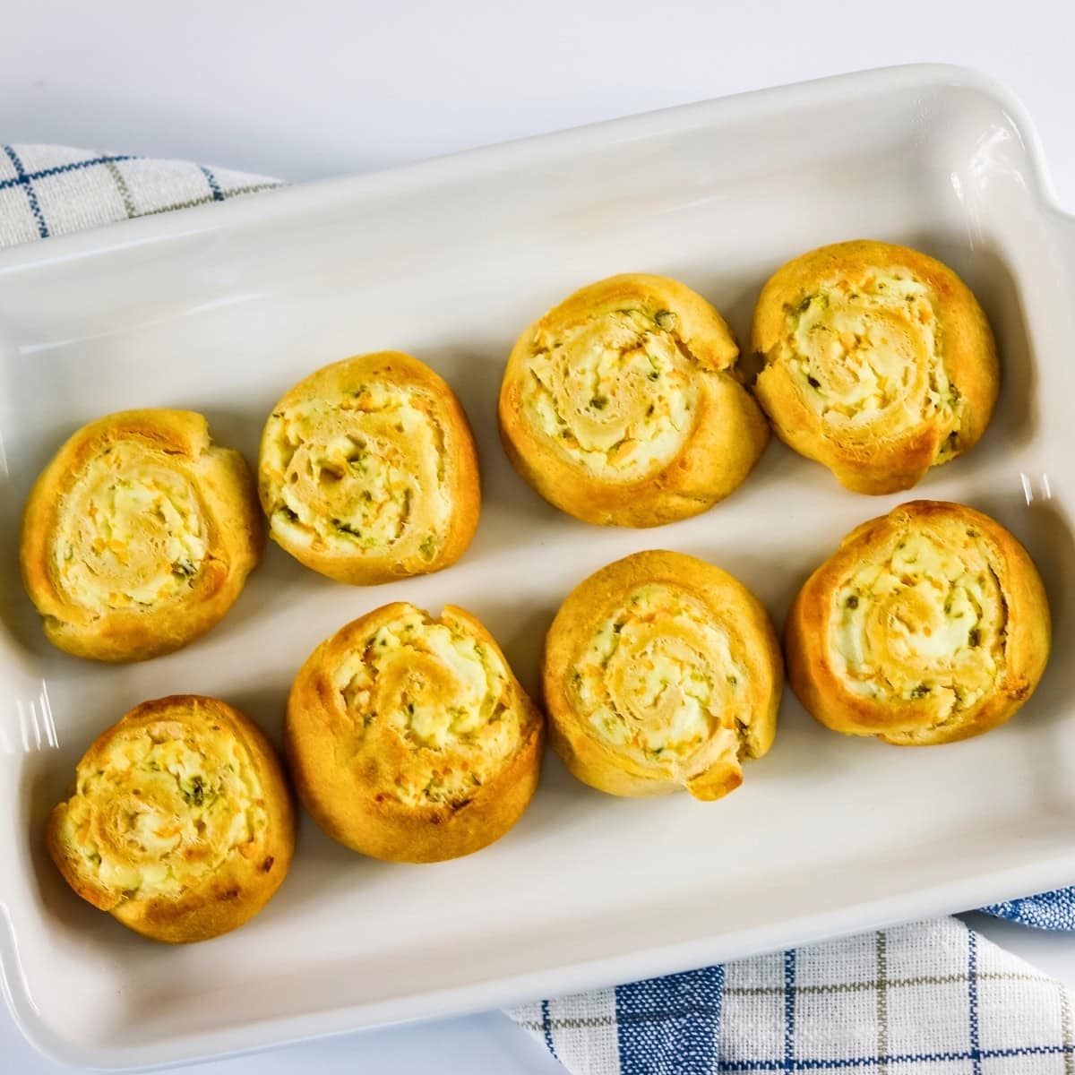 Eight golden pinwheel pastries arranged in a white baking dish on a checkered cloth.