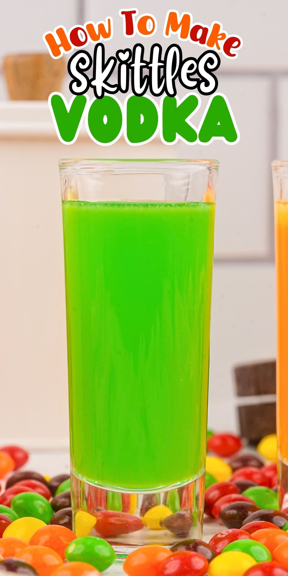A tall glass of bright green Skittles vodka surrounded by colorful Skittles.