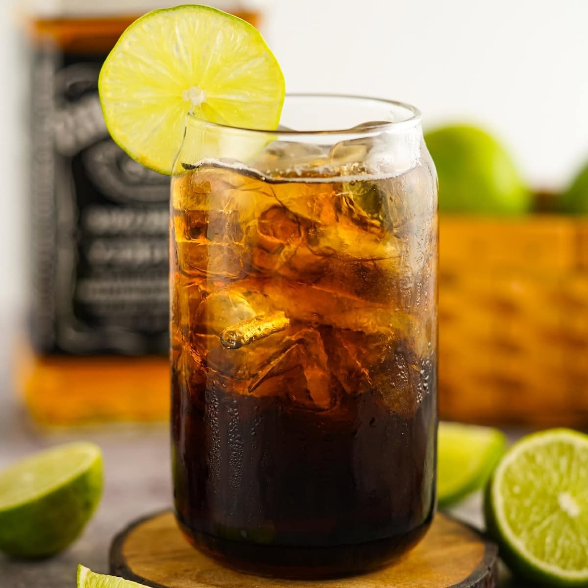 A glass of cola with ice and a lime slice garnish on a wooden coaster.