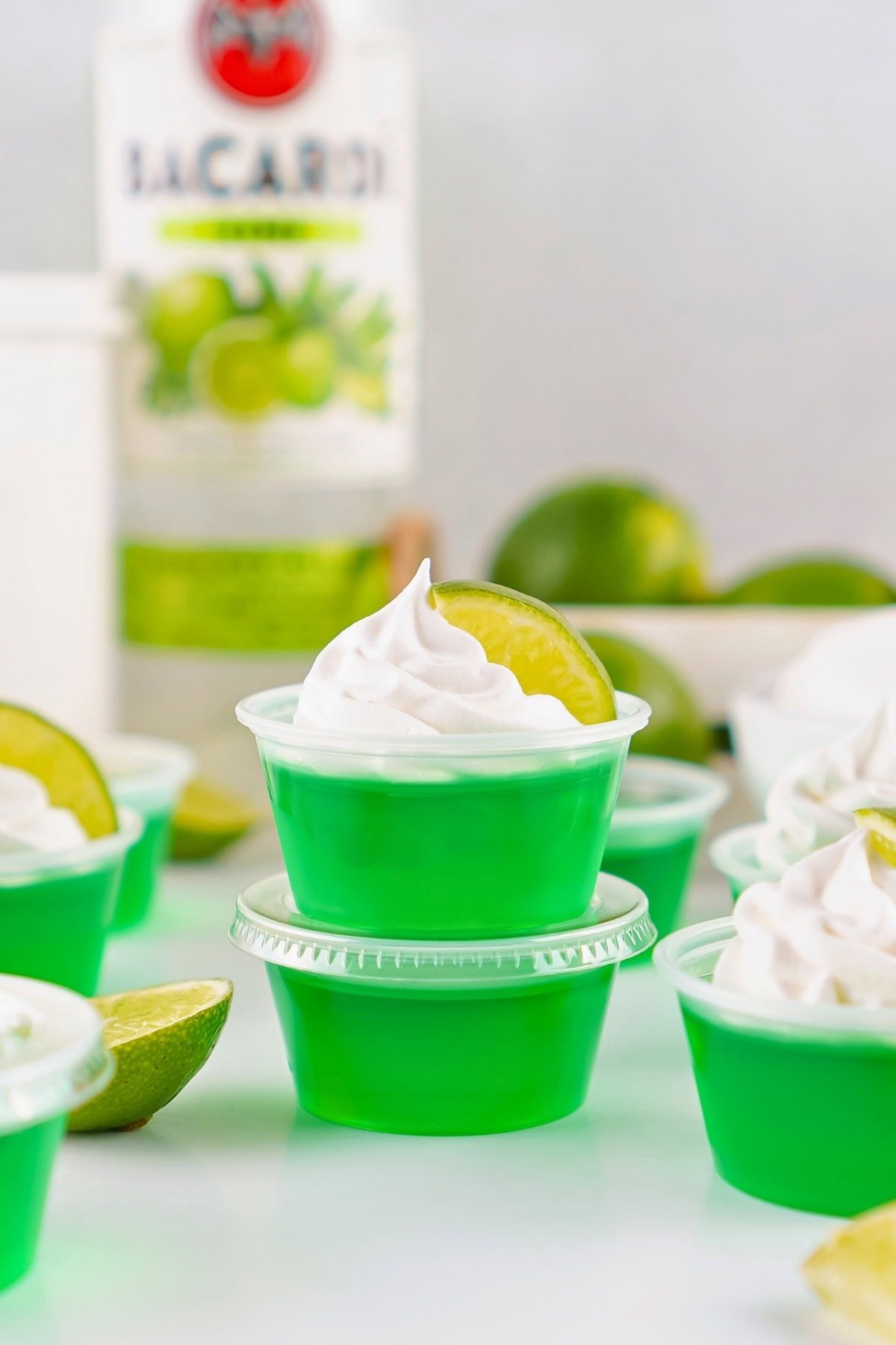 Green gelatin shots topped with whipped cream and lime slices, blurred rum bottle behind.