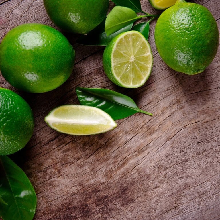 Fresh whole and sliced limes with green leaves on a rustic wooden surface.