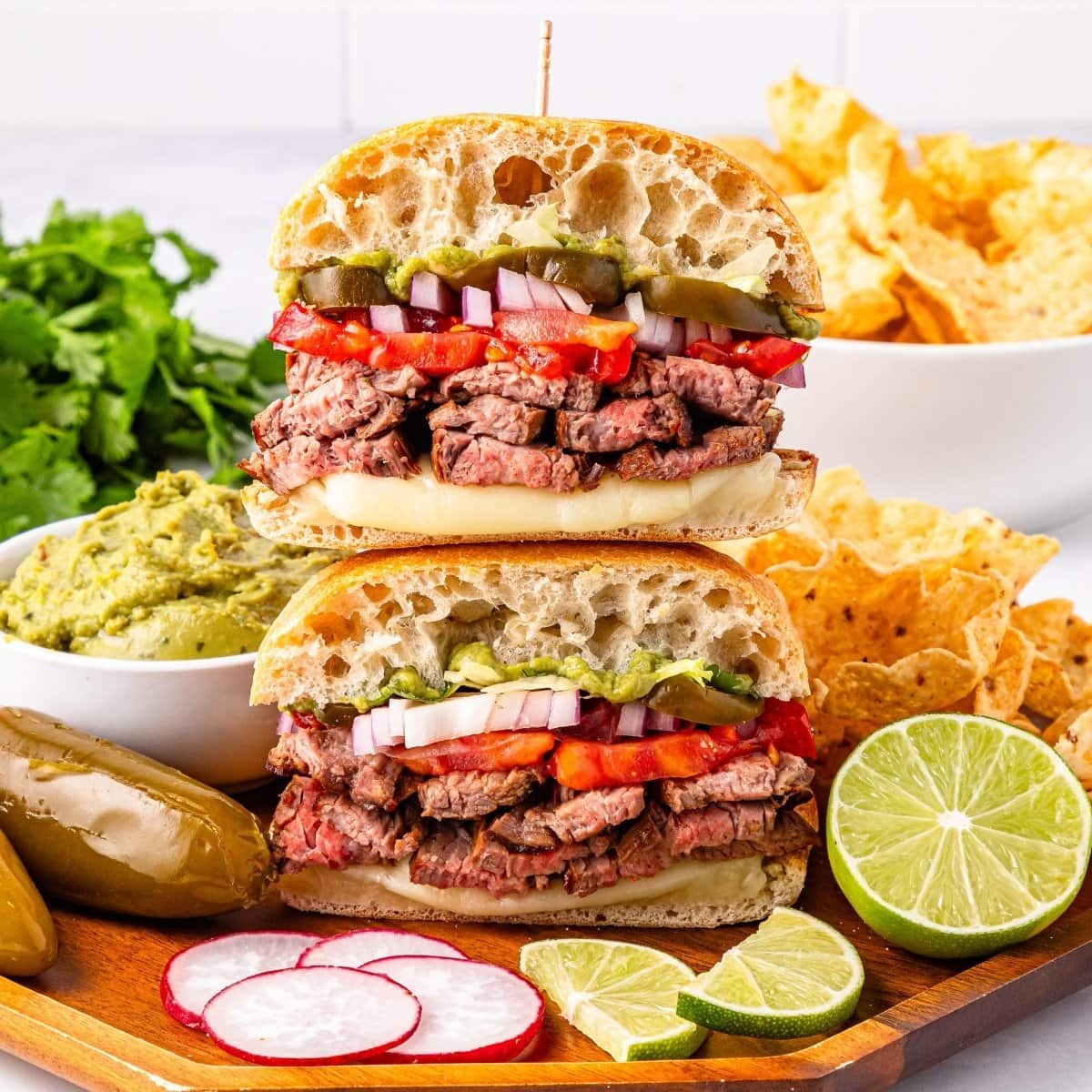 Stacked steak sandwiches with vegetables, chips, guacamole, limes, and jalapeños.