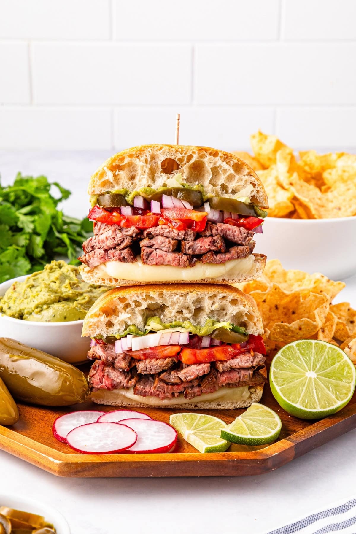 Two stacked steak sandwiches with chips, guacamole, radishes, and lime on the side.
