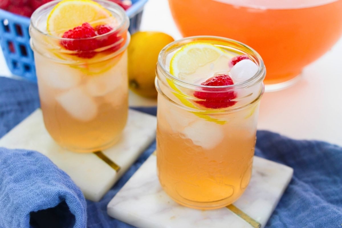 Two iced drinks with lemon slices and raspberries in mason jars on marble coasters.