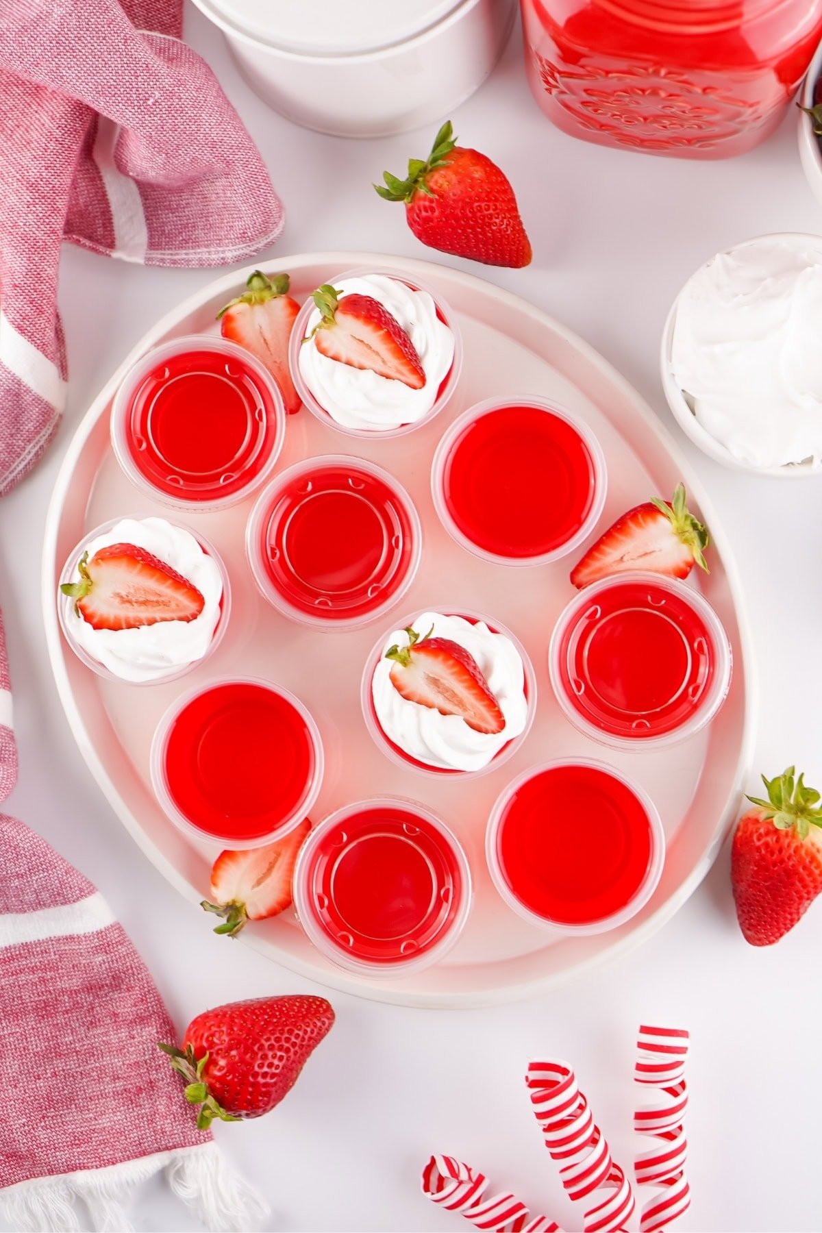 Red gelatin shots topped with whipped cream and strawberries on a white plate.