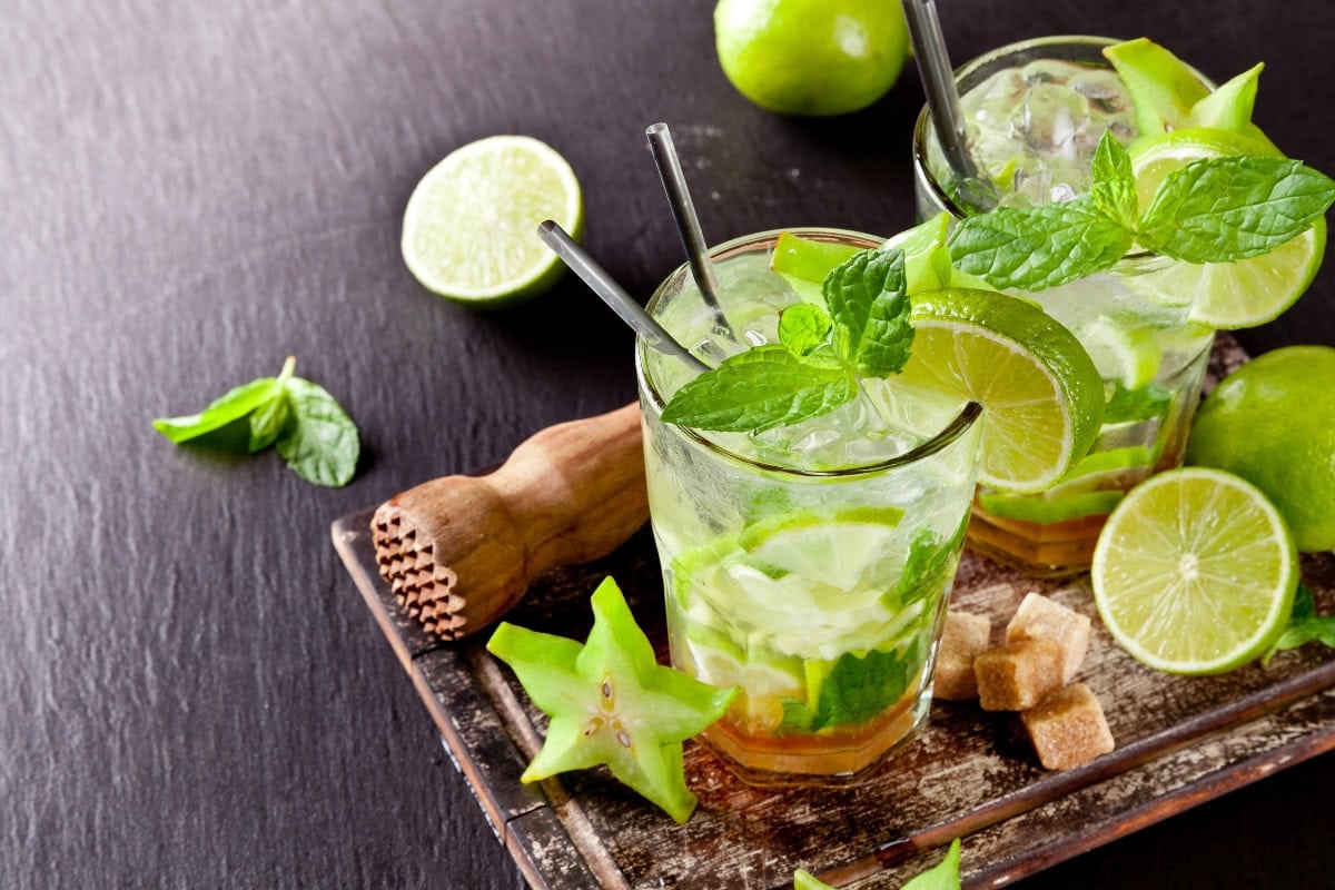 Two mojito cocktails with lime, mint, ice, and starfruit on a wooden board.