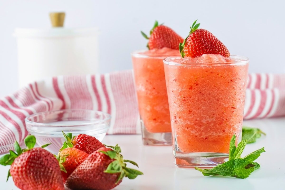 Two glasses of strawberry slush topped with strawberries, with fresh strawberries nearby.