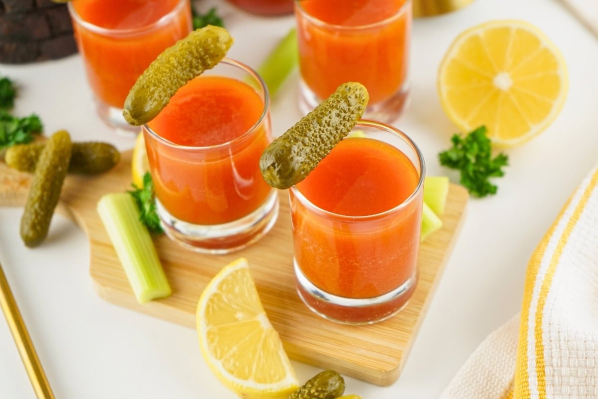 Three tomato juice shots garnished with pickles, lemon wedges, and celery nearby.