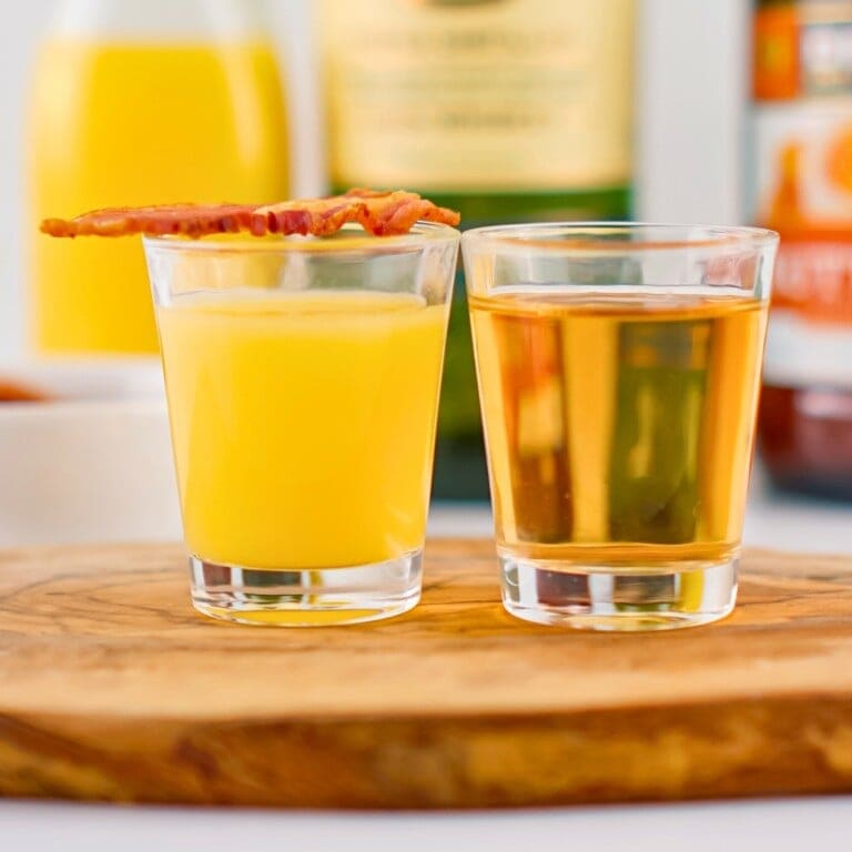 Two shot glasses, one with orange juice and one with whiskey, on a wooden board.
