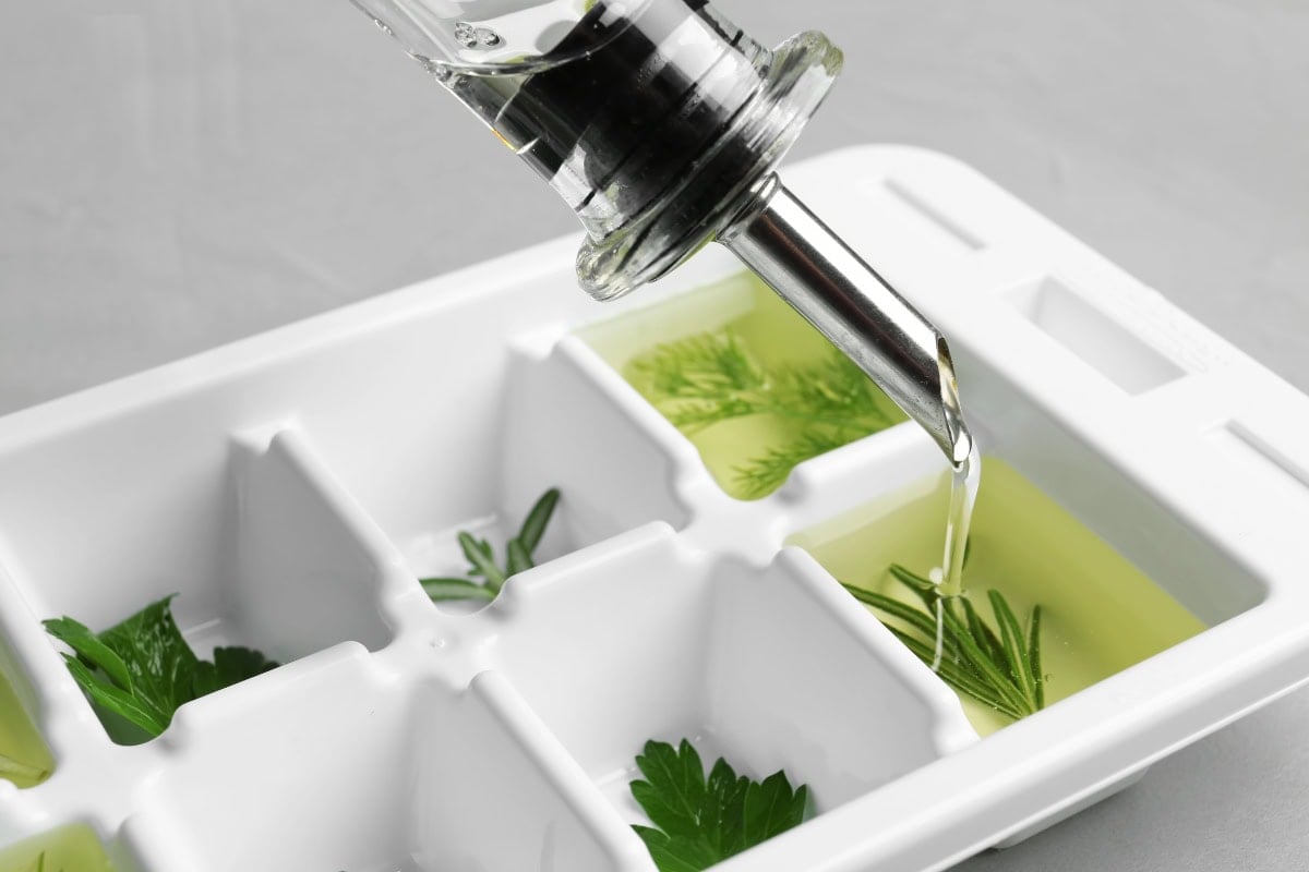 Pouring oil into an ice cube tray with fresh herbs inside each compartment.