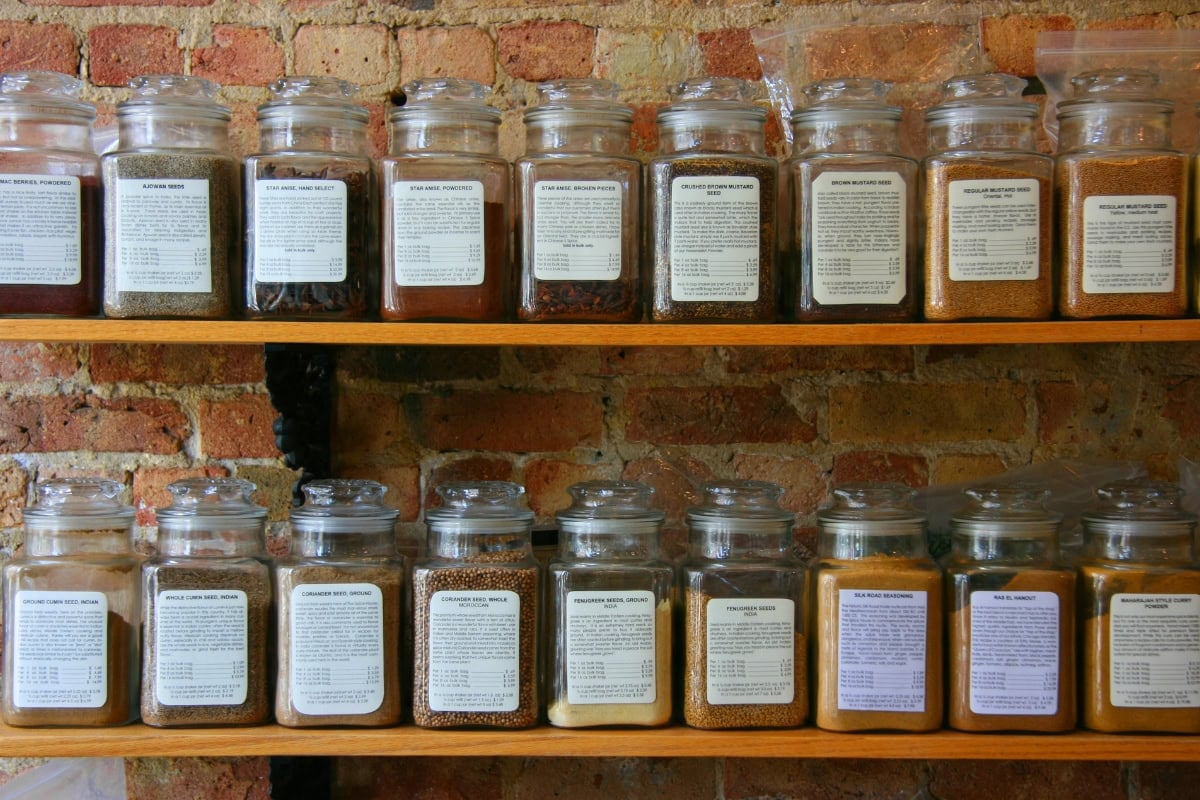 Glass jars filled with spices and grains lined up on wooden shelves against a brick wall.