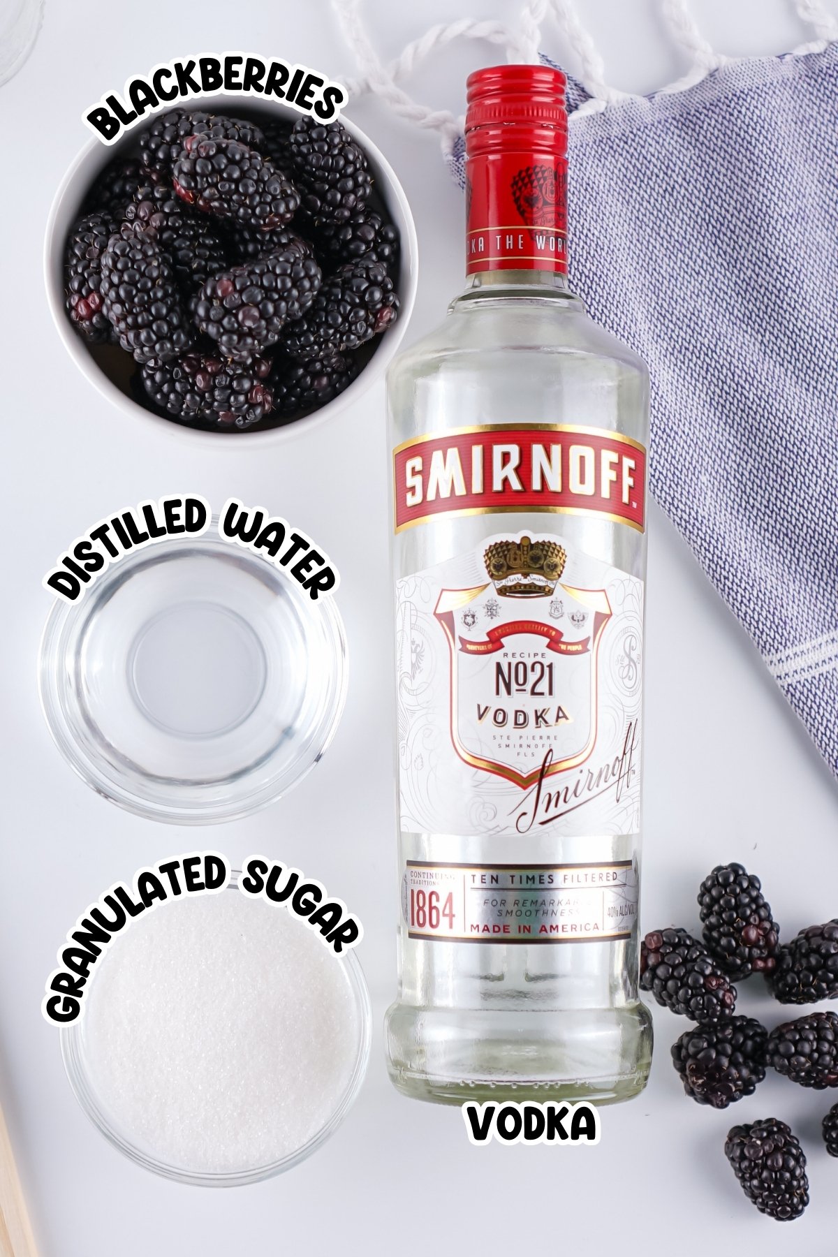 A bottle of vodka, blackberries, sugar, and water on a white surface.