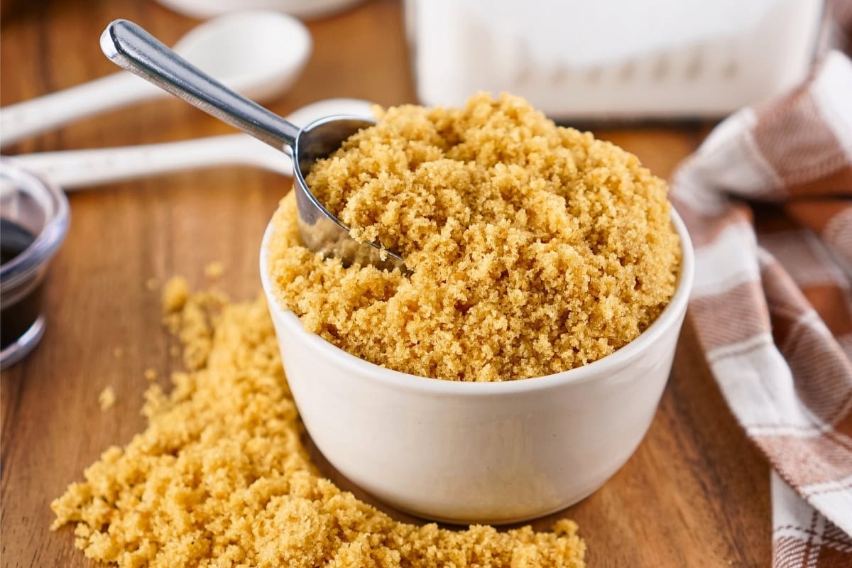 A bowl of brown sugar with a metal scoop and some sugar spilled on the table.