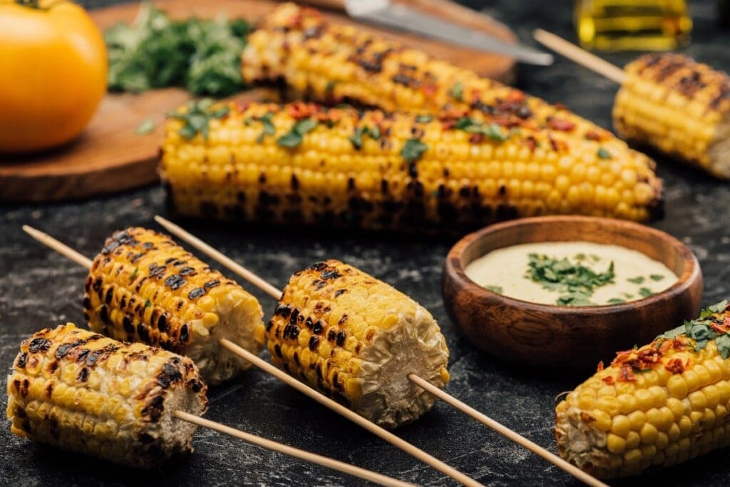 Grilled corn on the cob with herbs and dipping sauce on a dark surface.