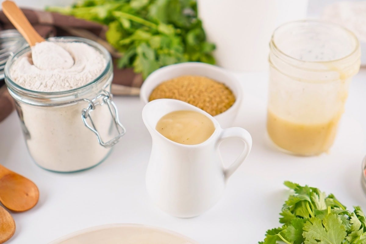 A white pitcher of gravy with flour, sugar, and herbs on a white surface.