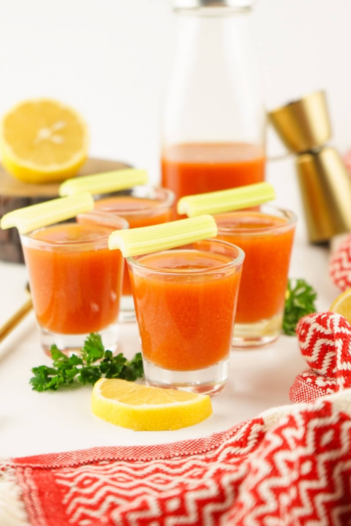 Four tomato juice shots with celery sticks, lemon wedges, and red patterned cloth.