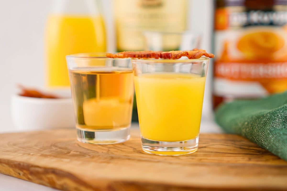 Two shot glasses, one with orange liquid and bacon, on a wooden board.
