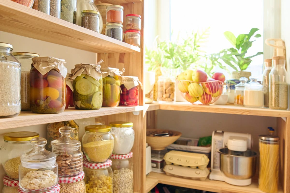 Wooden pantry shelves with jars of preserved food, pasta, and fresh fruits.