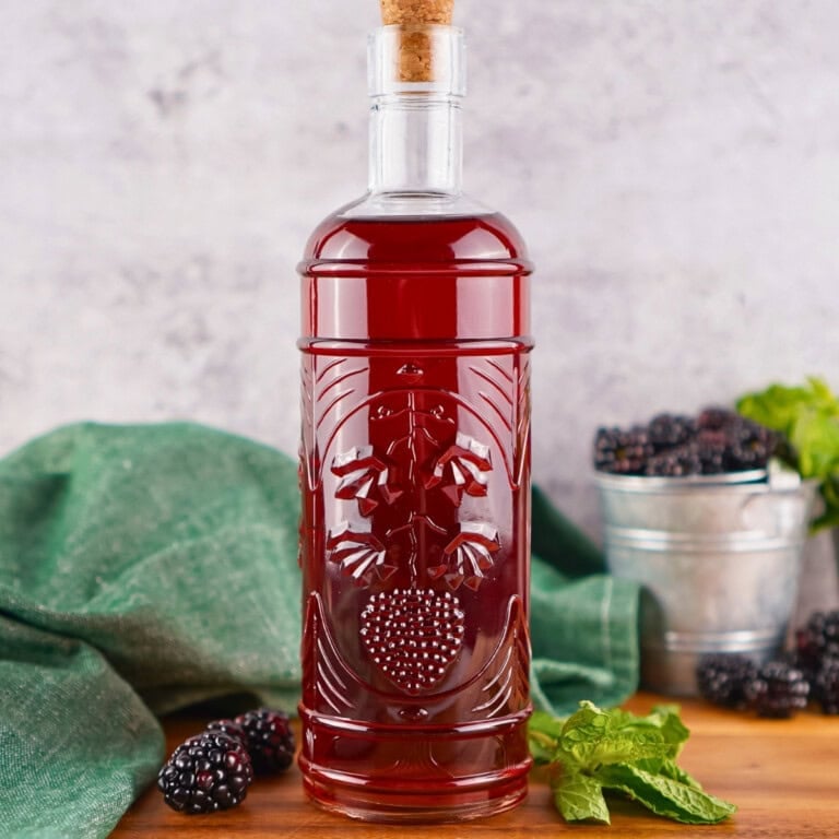 A glass bottle of blackberry liqueur with fresh blackberries and mint beside it.