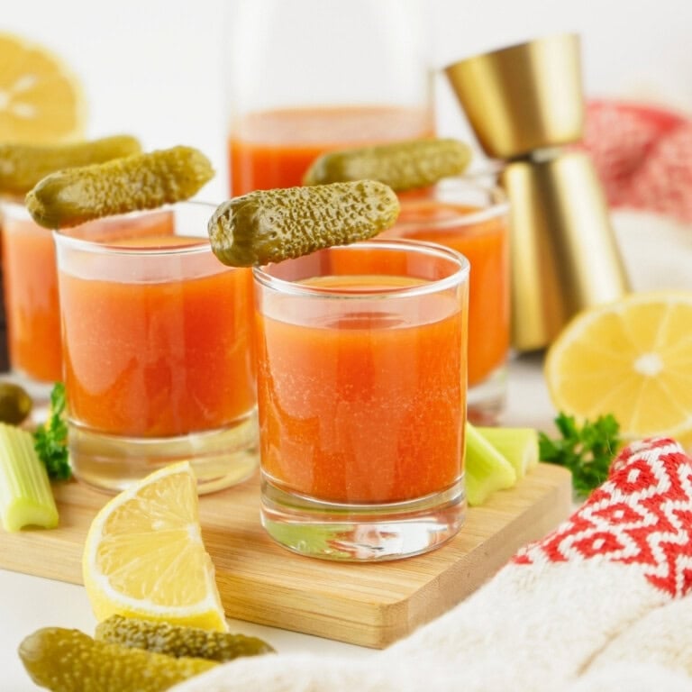 Three glasses of tomato juice garnished with pickles, surrounded by lemons and celery.
