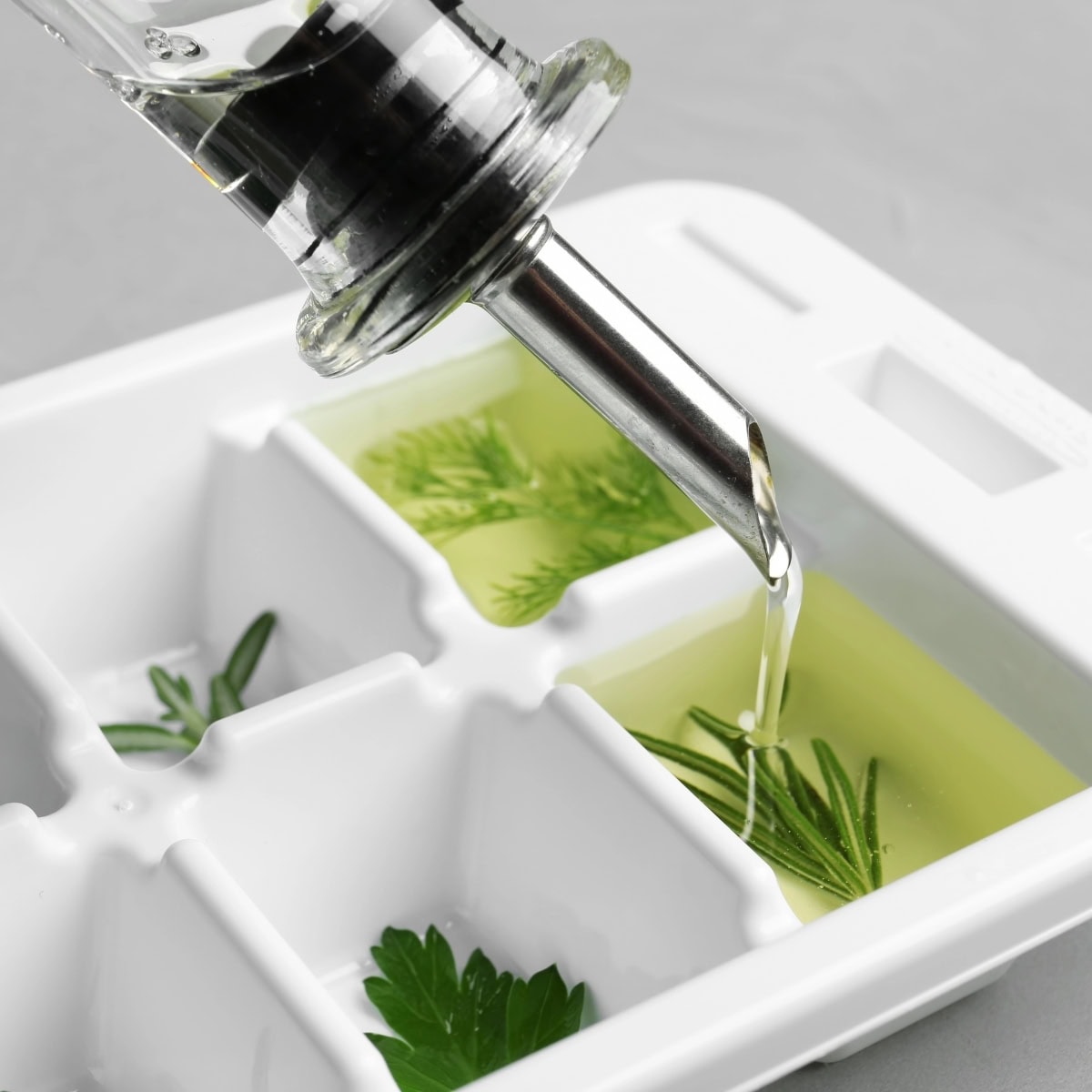 Olive oil being poured over herbs in an ice cube tray.
