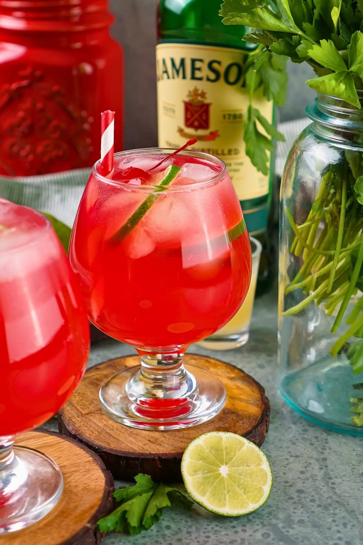 A red cocktail with ice, a straw, and garnish, beside lime and whiskey bottle.