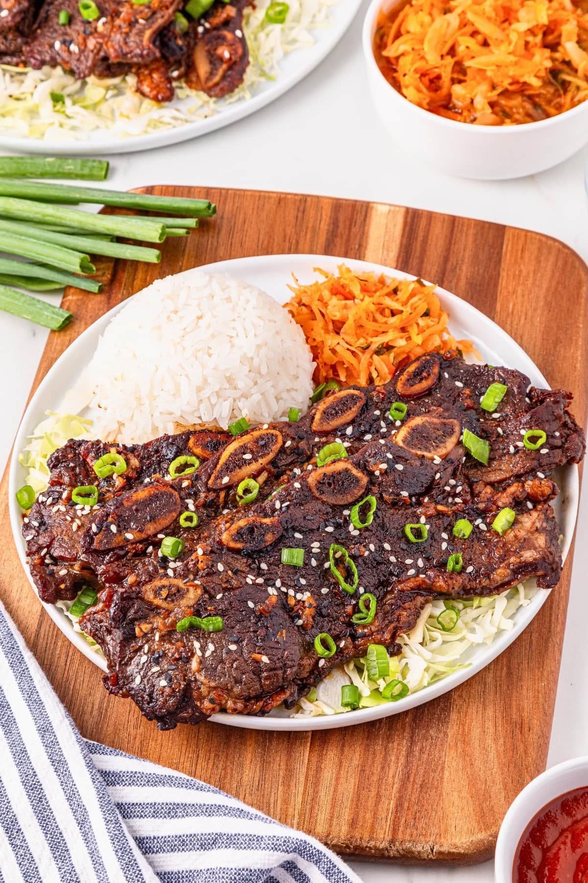 Plate of grilled beef short ribs, white rice, and shredded carrots, garnished with scallions.