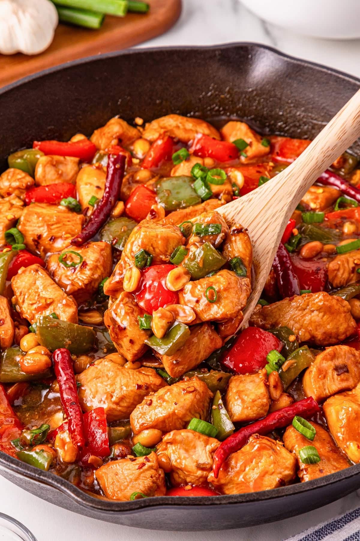 A skillet of kung pao chicken with peppers, peanuts, and chili peppers being stirred.