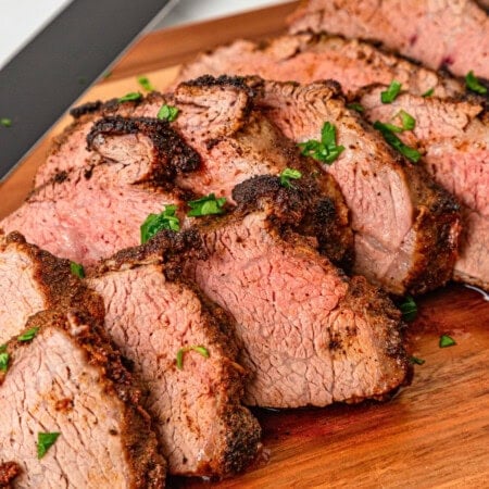 Sliced roast beef garnished with chopped herbs on a wooden cutting board.