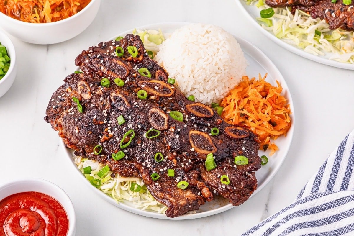 Plate of grilled short ribs with rice, shredded carrots, and green onions.