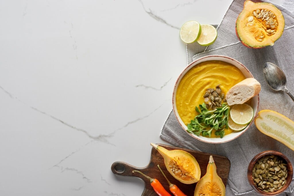 Bowl of pumpkin soup with bread, lime, seeds, and pumpkin on a table.