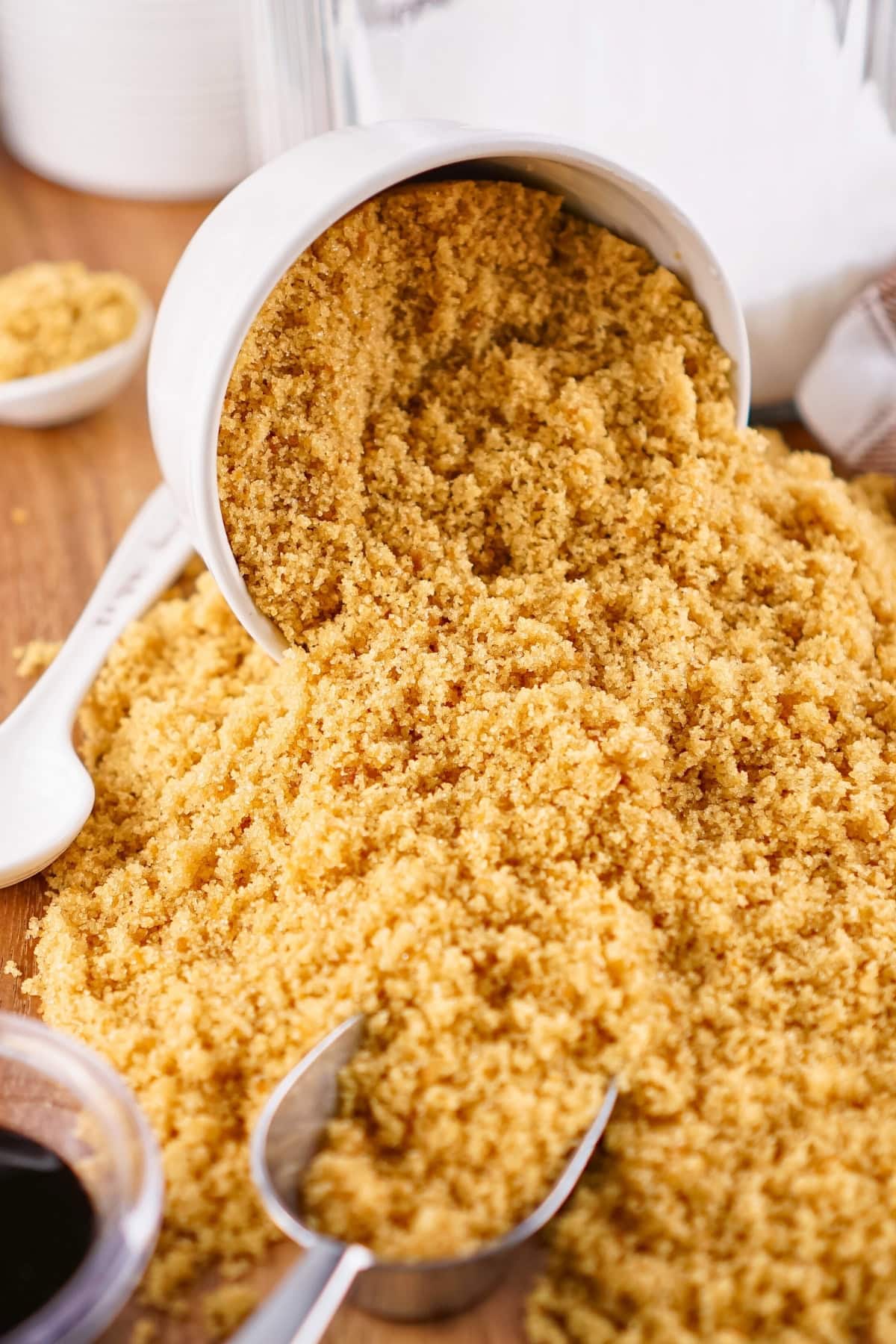 Brown sugar spilling from a white cup onto a wooden surface with a metal scoop.