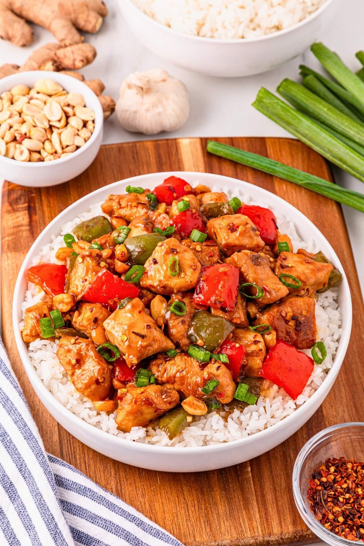 Bowl of kung pao chicken with rice, vegetables, and peanuts on a wooden board.