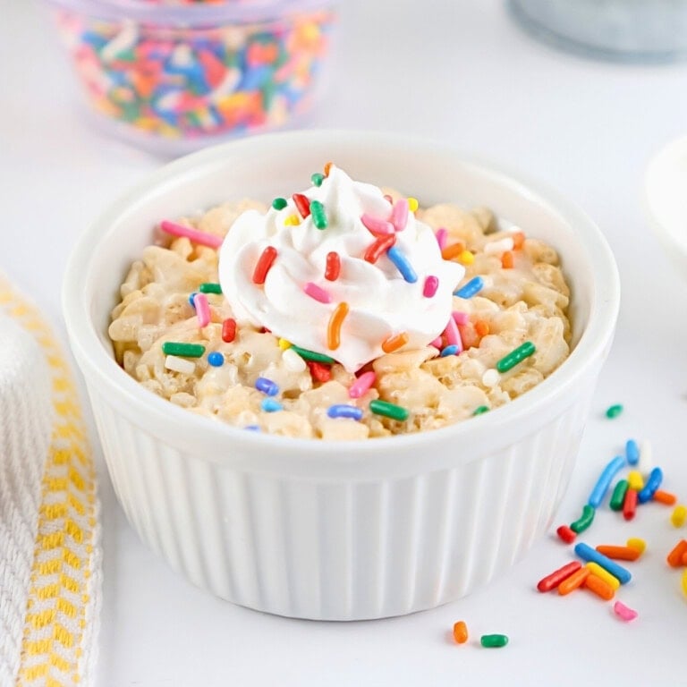 Rice Krispies treat in a white ramekin topped with whipped cream and rainbow sprinkles.