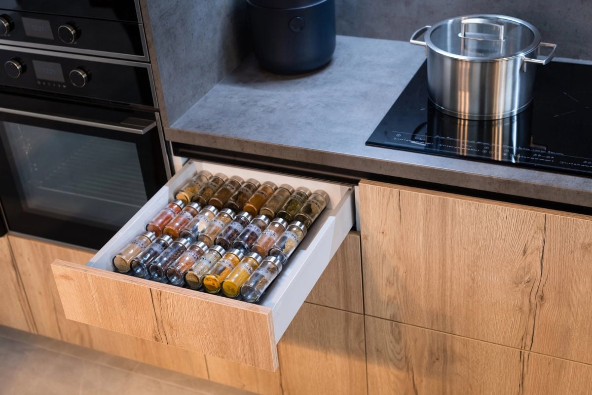 Open kitchen drawer with organized spice jars next to a stove with a pot on top.