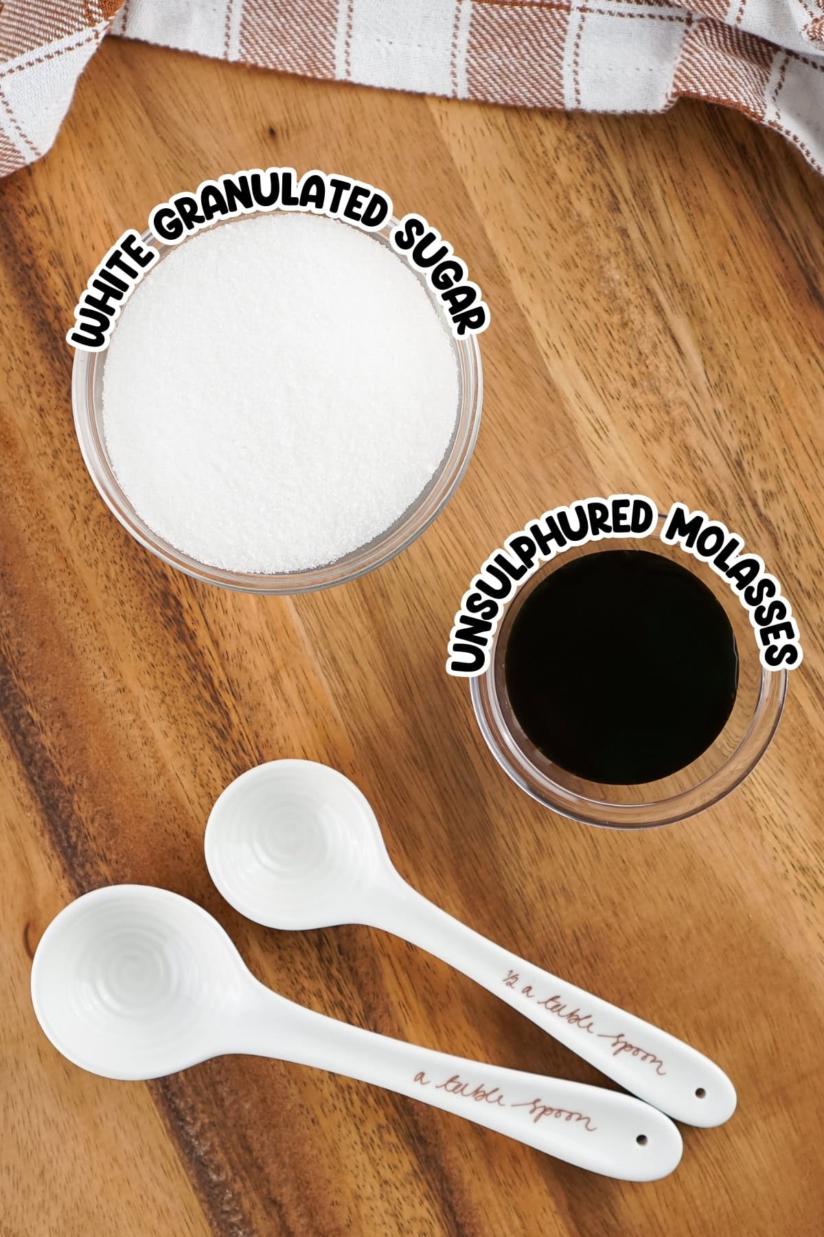 A bowl of sugar, a bowl of molasses, and two white measuring spoons on wood.