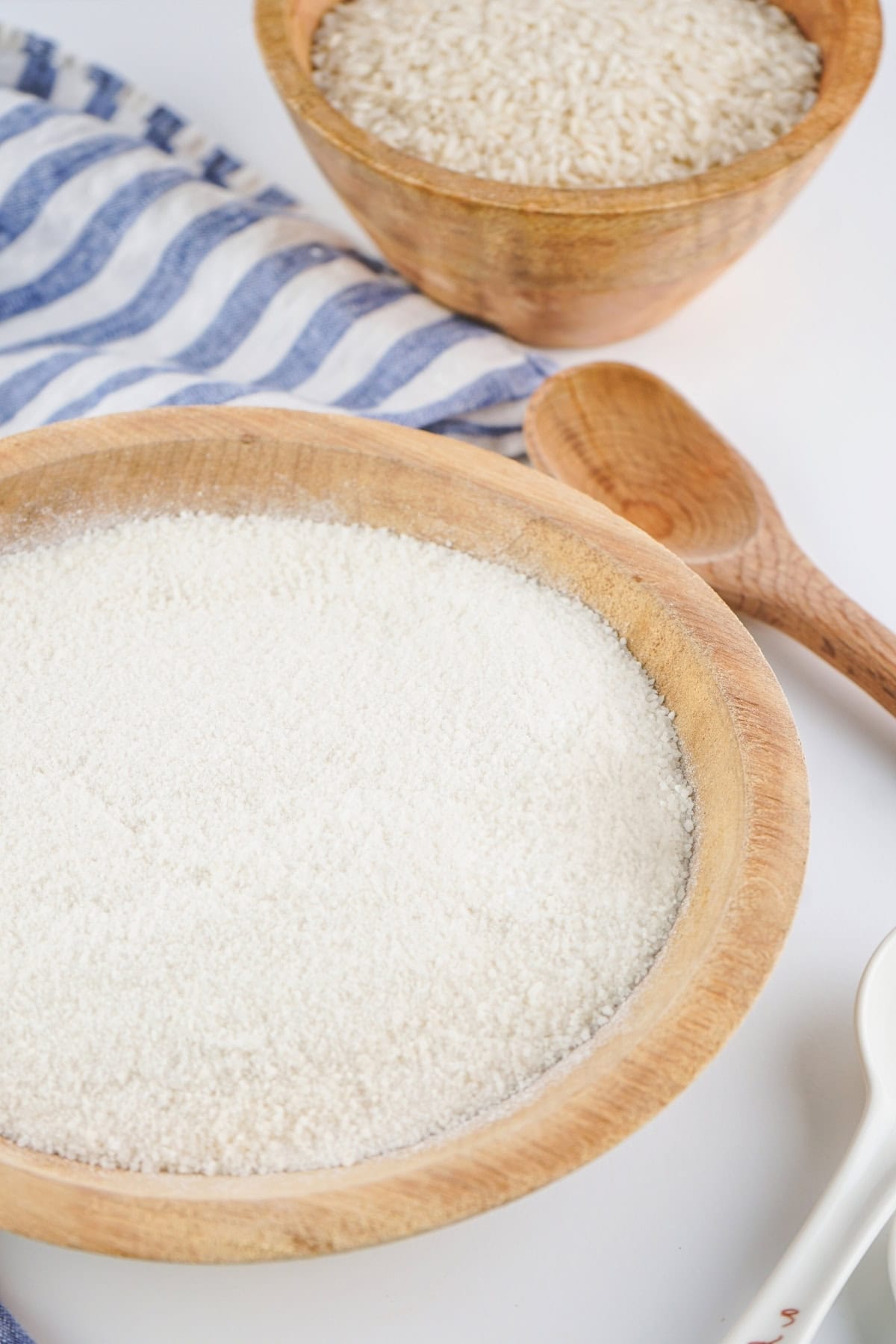 A wooden bowl filled with rice flour, with rice and a spoon in the background.