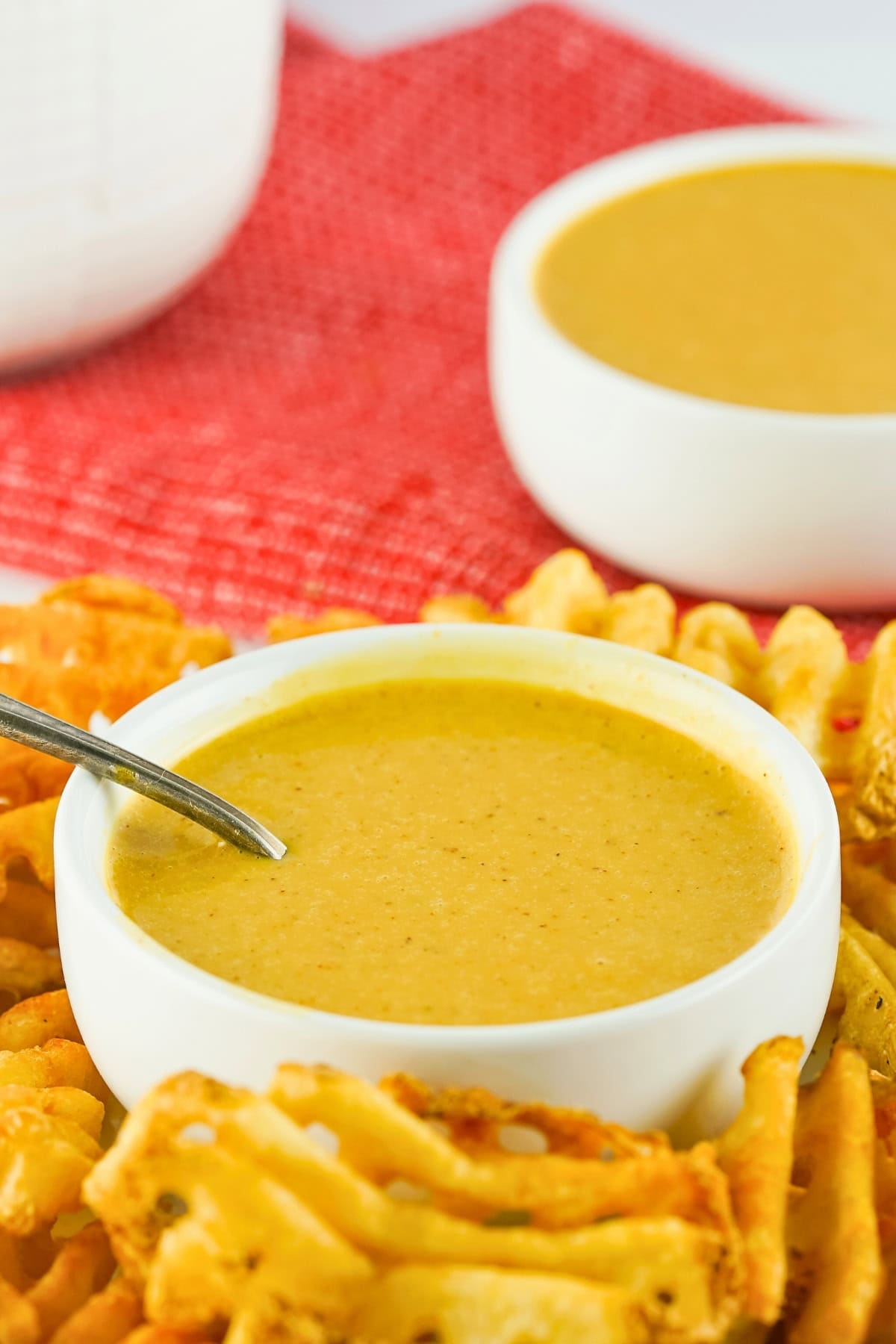 A bowl of cheese sauce with fries on a plate, set on a red textured cloth.