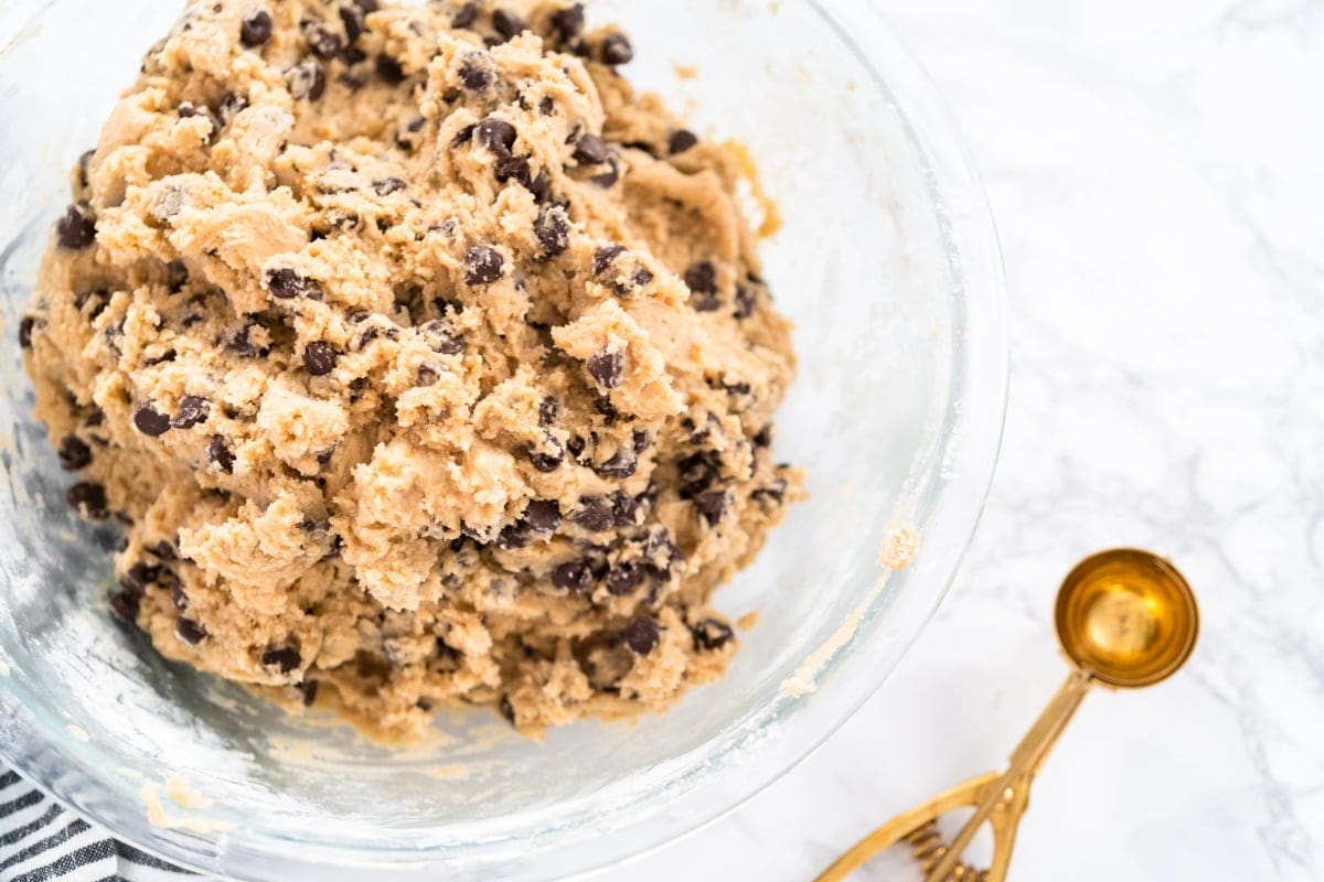 A glass bowl filled with chocolate chip cookie dough and a gold cookie scoop.