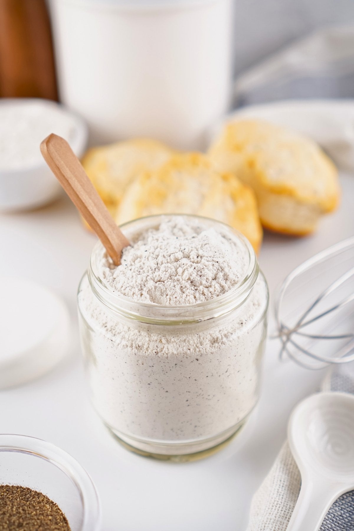 A jar of gravy mix with a small wooden spoon, biscuits and baking tools nearby.