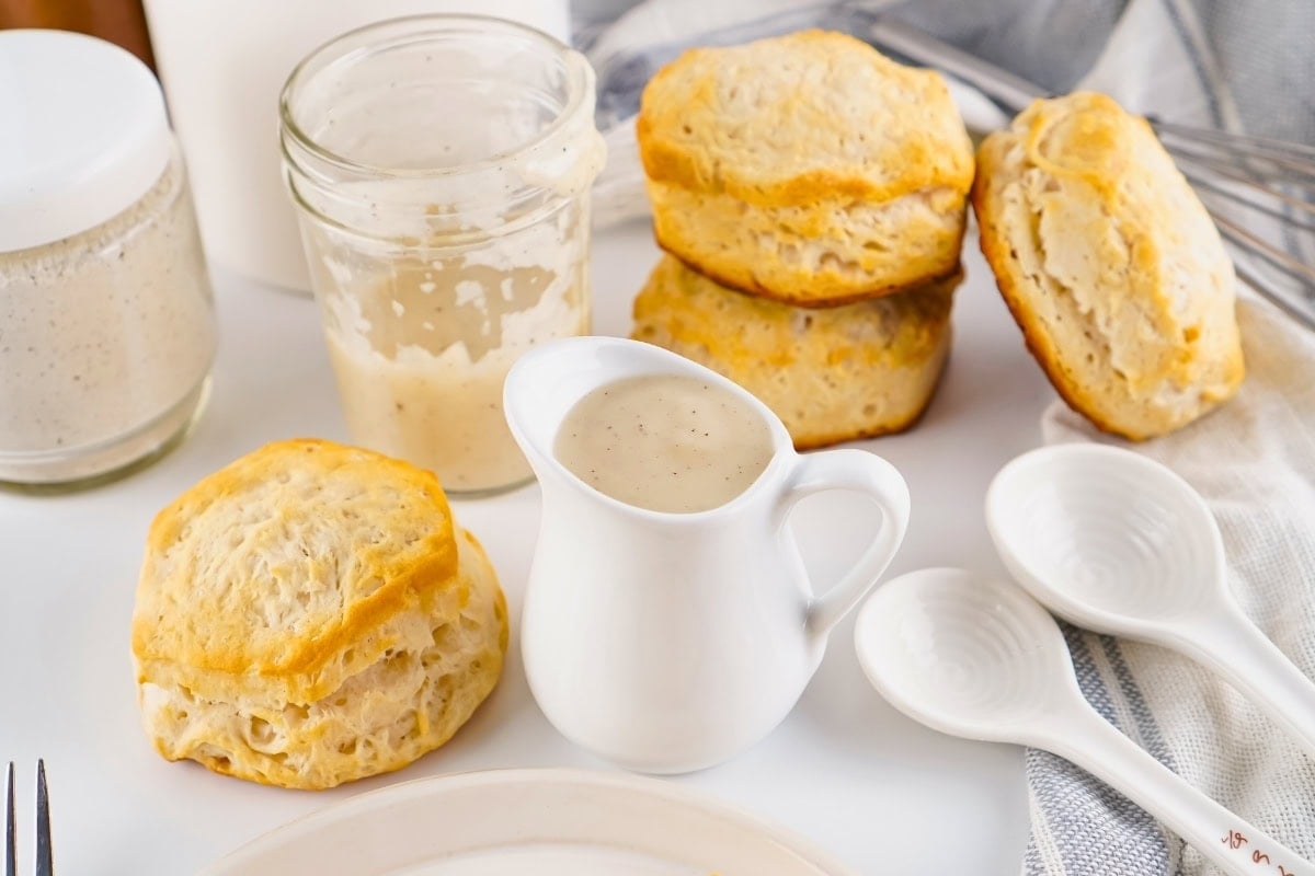 Biscuits with white gravy, jars, and spoons on a white table.