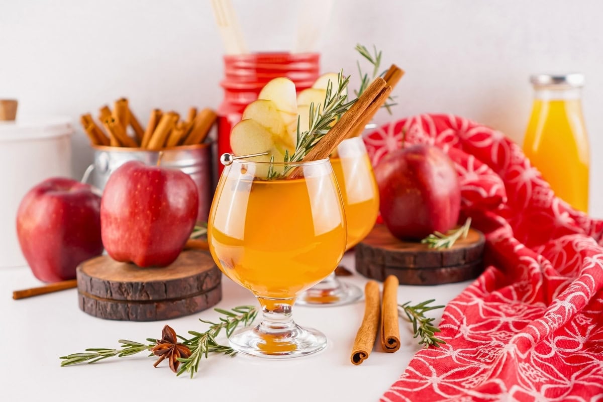 Apple cider in glasses garnished with apple slices, cinnamon, and rosemary.