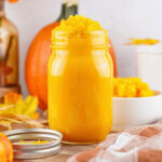 A glass jar of homemade pumpkin puree, surrounded by fresh orange pumpkins.