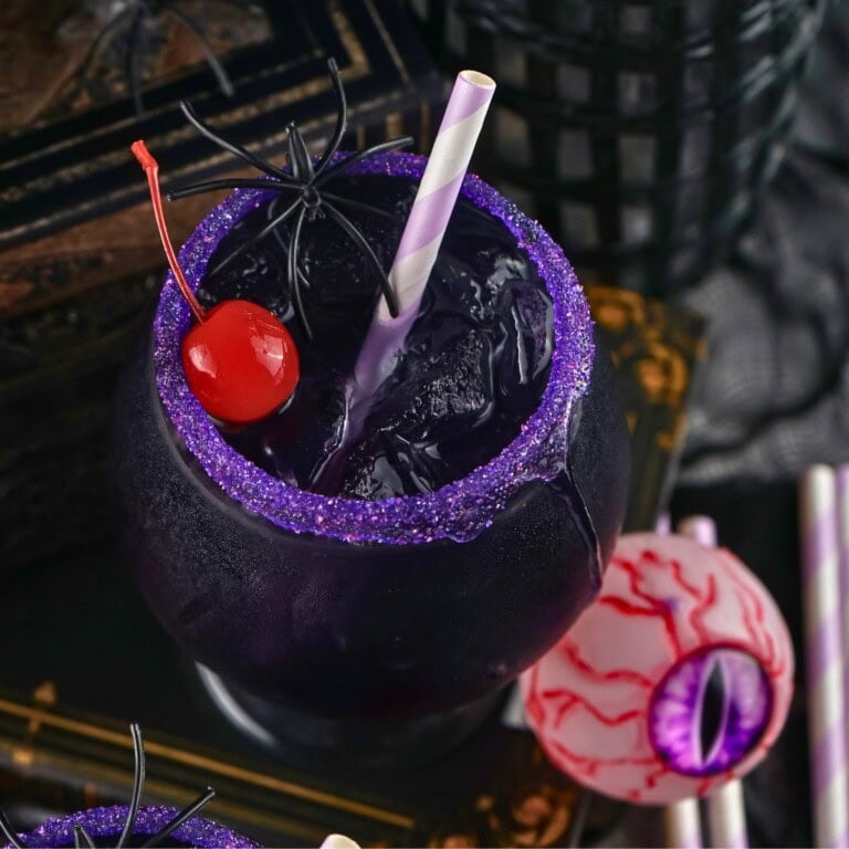A dark purple Halloween themed drink with a sugared rim, cherry, and spider.