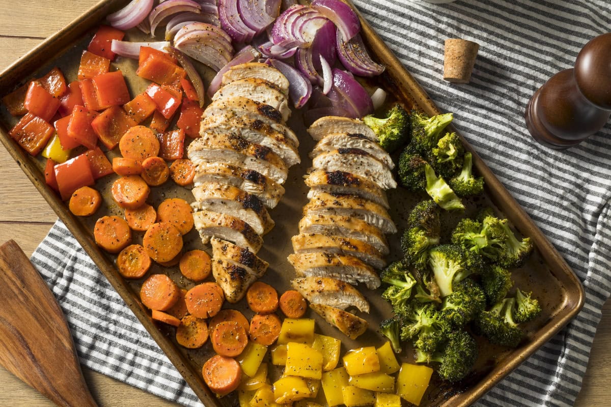 Sheet pan with sliced chicken breasts, broccoli, carrots, peppers, and red onions.