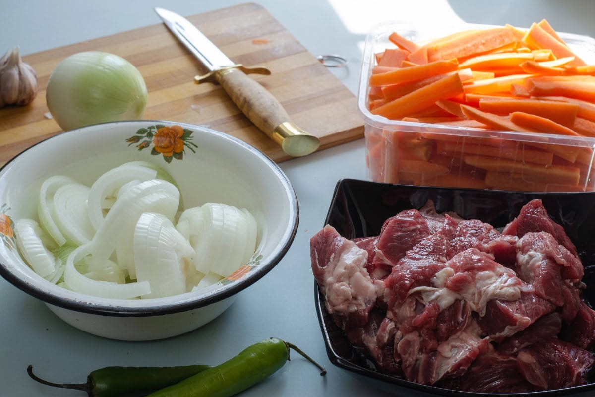 Sliced onion, raw meat, carrot sticks, chilies, and a cutting board with a knife.