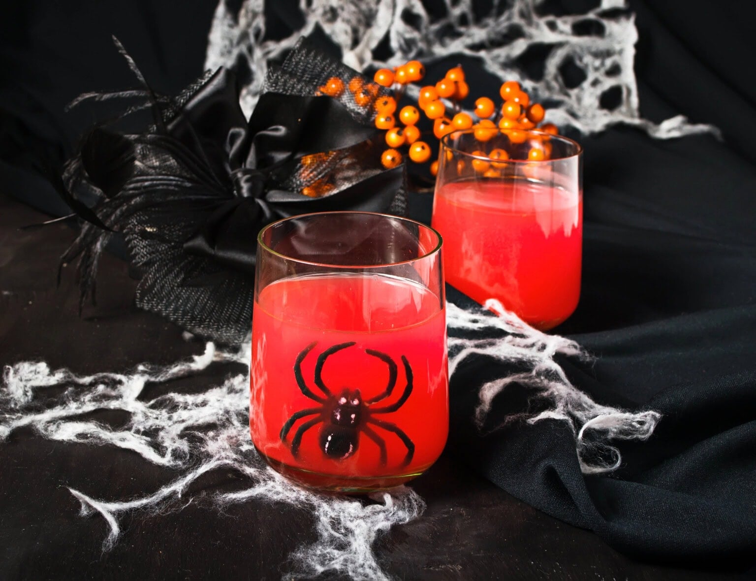 Two glasses of red drink with a spider decoration, surrounded by fake cobwebs.