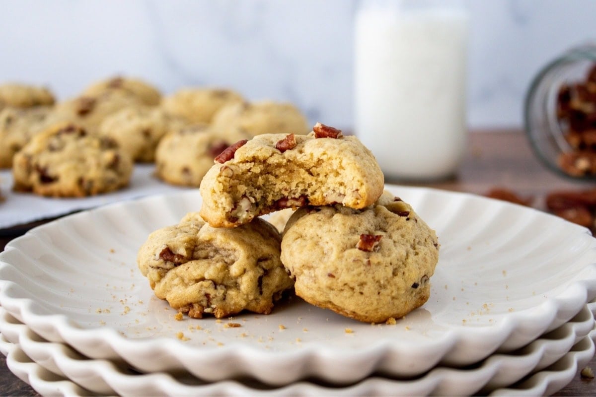 Three pecan cookies on stacked plates, one with a bite taken out, milk in background.